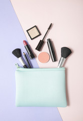Aerial view of make up products spilling out of a pastel blue cosmetics bag, on a pink and purple background