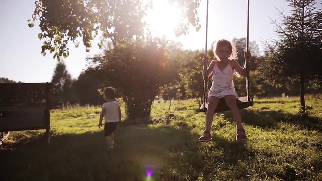 Beautiful cute brother and sister playing outdoor. Girl swinging, boy walking on the grass.