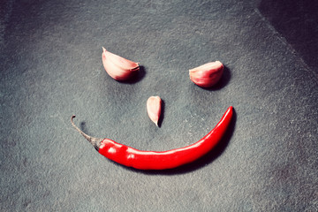 Smiling face made of red chili pepper and cloves of garlic on slate background, conceptual picture, selective focus.