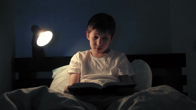 Boy resting in his bed in the evening and reading a book turns off light and goes to sleep