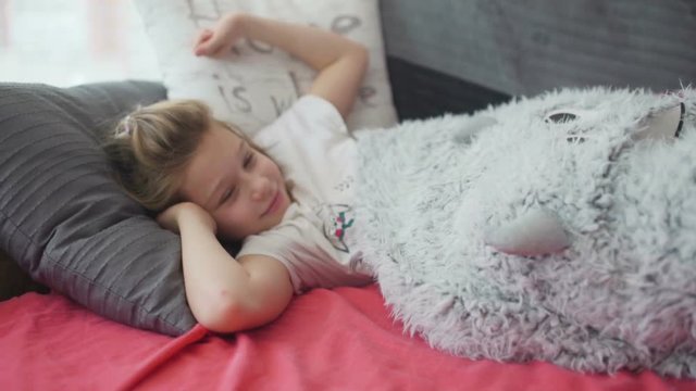 Cute little yawning girl in pajamas lying in a bed under a white blanket on sunny morning