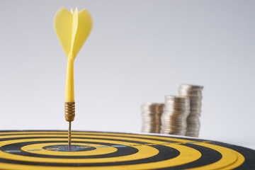 Yellow dart hit in the center of a target with stack of coins behind on white background with copy...