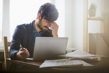 Portrait of exhausted young man sitting at workplace in modern office against window rubbing his forehead and closing eyes in pain while working at laptop
