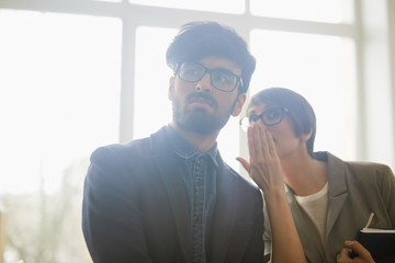 Portrait of two office workers gossiping about newcomer: young woman leaning in to whisper to mans ear as they both looking judgingly at someone