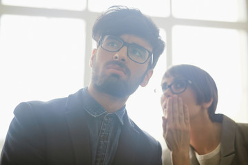 Closeup portrait of two office workers gossiping about newcomer: young woman leaning in to whisper...