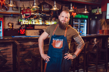 Fototapeta na wymiar Bearded barman with tattoos and watches wearing an apron standing near the bar.