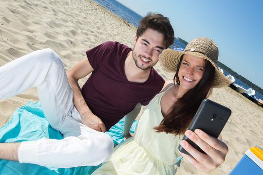 gorgeous young couple sitting at the beach and doing selfie