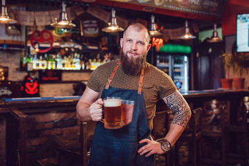 Bearded barman with tattoos wearing an apron standing near the bar and holding a glass of beer