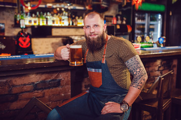 Bearded barman with tattoos wearing an apron sitting near the bar and holding a glass of beer