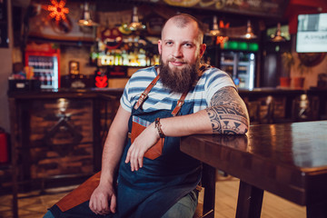 Bearded barman with tattoos wearing an apron sitting at the bar.