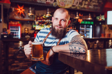 Bearded barman with tattoos wearing an apron sitting at the bar and holding a glass of beer.