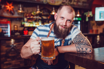 Bearded barman with tattoos wearing an apron sitting at the bar and holding a glass of beer.