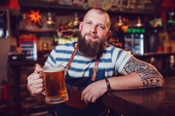 Bearded barman with tattoos wearing an apron sitting at the bar and holding a glass of beer.