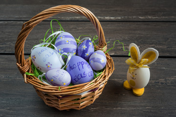 Easter wicker basket with colored eggs and a small egg rabbit on grey wooden board.