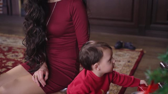 Beautiful and elegant young mother with long black hair sitting next to a Christmas tree. She poprovlyaet his little son sweater with deer. Malik sits nearby and playing with toys