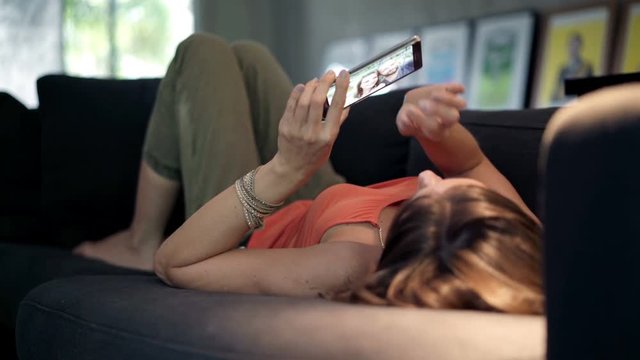 Young woman lying on sofa and browsing photos on smartphone, 4K
