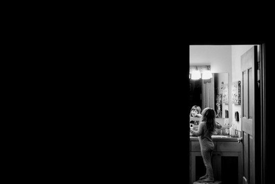 Young girl standing on stool in bathroom, brushing teeth, looking in mirror, black and white