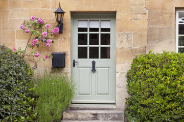 Naklejka premium Light green wooden doors in an old traditional English lime stone cottage surrounded by climbing pink roses, lavender, on summer day