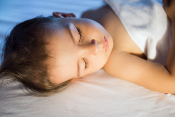 Close-up portrait of adorable baby girl sleeping in white bed at night. Little toddler taking a nap in dark room in lamplight. Bed time for kids. Bedroom and nursery interior.