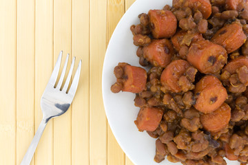 Top close view of beans and hot dogs on a plate with a fork to the side atop a wood place mat.