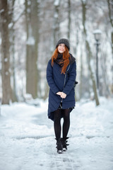Beautiful winter portrait of young adorable redhead woman in cute knitted hat winter having fun on snowy park path
