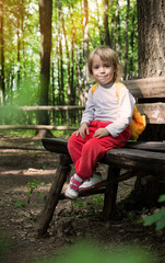 Naklejka na ściany i meble Little girl sitting on a bench in the park in the open air in anticipation of camping. Travel and adventure with your child. Cute little traveler with a backpack