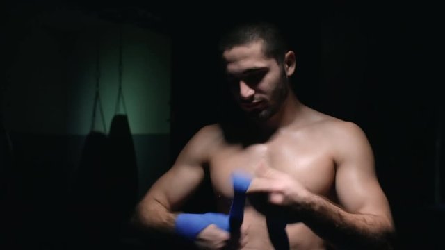 Muscular man standing in dark ring and wrapping hand in protective band before beginning of workout
