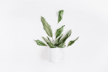 Blank template of white mug and green leaves bouquet on white background. Flat lay, top view.