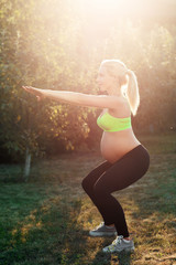 Pregnant woman squatting outdoor, sunrise light. Side view on expectant female in sportswear...
