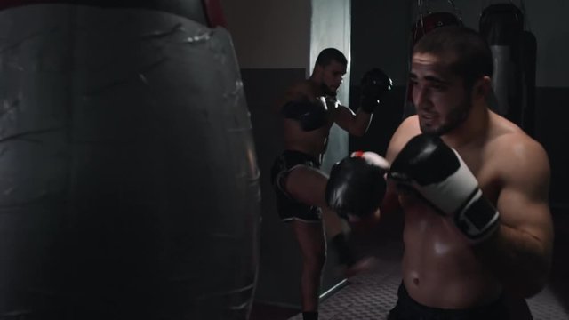Two muscular shirtless MMA fighters in boxing gloves punching bags in dark gym in slow motion