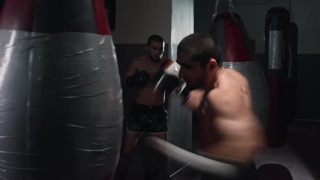 Two professional MMA fighters in boxing gloves punching bags in gym 