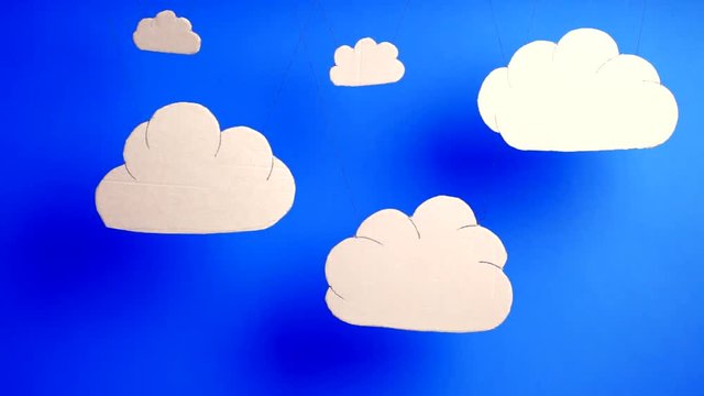 Moving white cloud cardboard cutouts hanging in front of blue sky background. Cartoon style weather concept of a fine breezy day