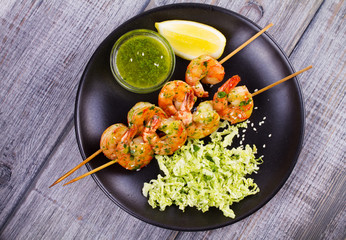 Cilantro grilled shrimps on skewers with sesame napa cabbage, green butter sauce and lemon. Skewered prawns on black plate. View from above, top studio shot