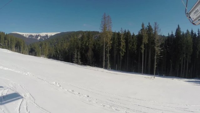 Ski lift ride clip, side view, with snow covered ski trails below, mountains and trees in the background and vacant chairs passing by
