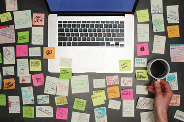 Top view on messy office desk with laptop, coffee and post it notes all around. - obrazy, fototapety, plakaty