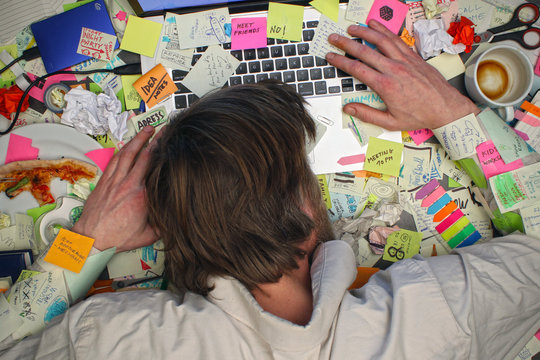 Overworked man sleeping on laptop computer with post it notes all around his office desk.