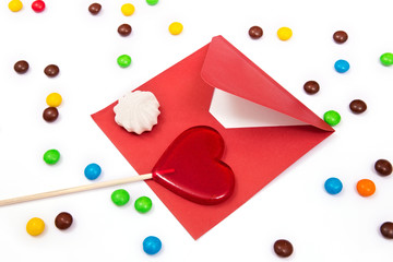 Gift for Valentines day. Red envelope, meringue, and red lollipop in the shape of a heart on the white background with colorful button-shaped chocolates.