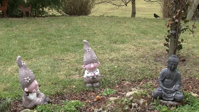 Im Vordergrund drei Gartenfiguren und im Hintergrund l&auml;uft bei leichtem Schneefall ein Amselm&auml;nnchen (Totale)