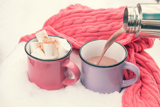 Pouring hot chocolate in the cup. Hot chocolate with marshmallow in pink and violet two cups wrapped in a cozy winter pink scarf on the snow-covered table in the garden. Coloring and processing photo