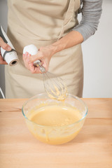 adult woman with beige apron, after whipping cream in glass bowl, with whisk piece in hand of electric mixer, to cook a sponge cake
