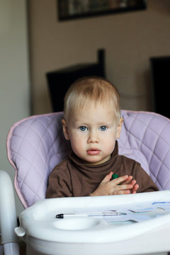 Little blonde ordinary baby boy sitting at chair for feeding and draws