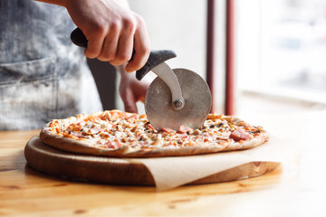 Closeup hand of chef baker in uniform blue apron cutting pizza at kitchen