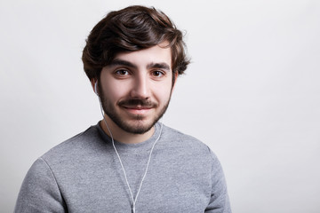 Close up portrait of stylish hipster with beard looking pensive, listening to music online on white earphones using some electronic gadget  looking happily directly into the camera