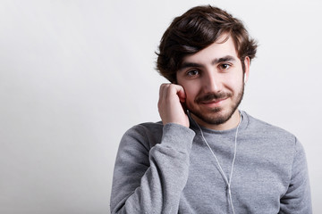 Young hipster with big dark eyes modern hairstyle and beard wearing casual grey sweater listening to the music or audiobook with his earphones holding his hand on earphones having happy expression