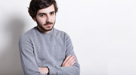 Headshot of fashionable young hipster with trendy hairstyle and thick beard dressed in grey sweater keeping his arms crossed having tires and thoughtful expression. Confident man with crossed hands