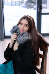 Beautiful young girl with long dark hair smiling sitting in a fur coat with a fur collar silver fox. She looks straight into the camera against the window.