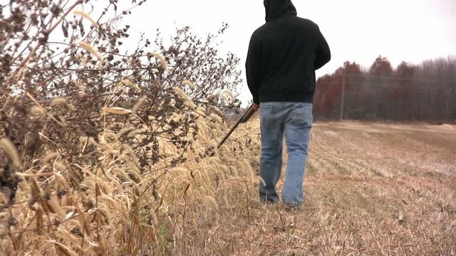 A man holding a 20 gauge shotgun is walking through a field looking for rabbit.
