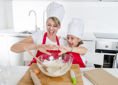 young mother and little sweet daughter in cook hat and apron cooking together baking at home kitchen