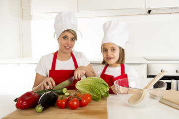 mother and little daughter cooking together with hat apron preparing salad at home kitchen