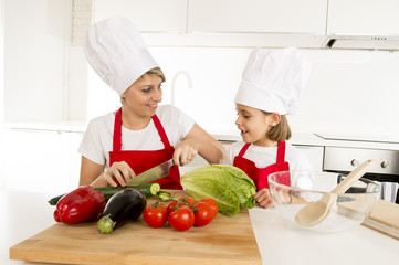 mother and little daughter cooking together with hat apron preparing salad at home kitchen